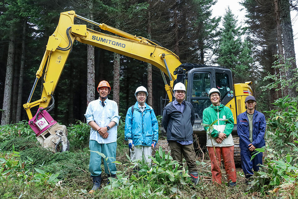 高性能林業機械フェラーバンチャ（立木を切り倒し、そのまま掴んで集積する機械）と天塩研究林の職員たち。中央が高木研究林長。右から2番目が藤田 達也 技術専門職員
