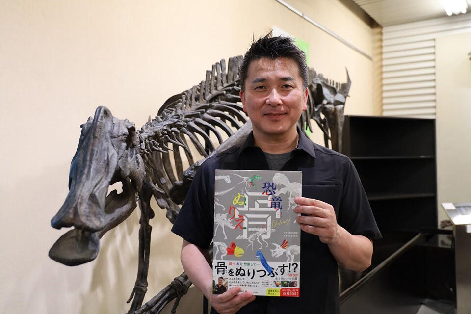 『恐竜 骨ぬりえ』を手にする小林教授本学総合博物館内に展示されているニッポノサウルスの骨格標本とともに